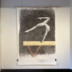 Large scale charcoal drawing of gymnast balancing on hands with back arched and legs split, with one leg straight and one leg bent. Blue collaged triangles near foot and and chest showing effort and force on the body, and ared collage dot near back of gymnast representing centre of gravity. The figure is on a gymnastics beam that is beige in colour with red leg beneath.
