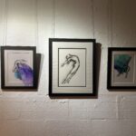 Three black framed figure drawings on a white wall. The central piece is larger, and the pieces on the left and right feature translucent ink ovelays in blend of blue, green, purple.
