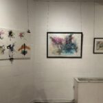 Three artworks on a white wall. From left to right: six mixed media mark-making exercises on white paper, hung with bulldog clips; a drawing of a figure at different stages an aerial skill againt a pink, blue and beige abstract background, in a black frame; a smal charcoal drawing of figure leaping against a pink, blue and yellow abstract background, in a black frame.